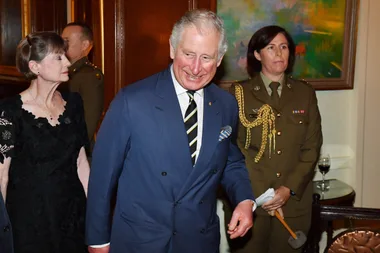 Man in a blue suit smiling at a formal event, accompanied by a woman in black and a military officer in uniform.