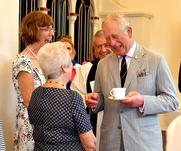 Prince Charles laughing and holding a teacup while engaging with a group of people in a social setting.