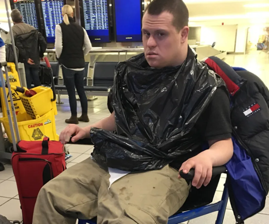 A teenager with Down syndrome sits in a wheelchair at an airport, wearing a black plastic cover.