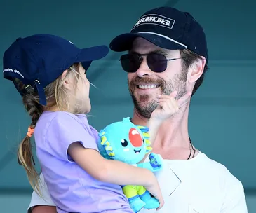 Father in cap and sunglasses holds daughter with stuffed toy at event, both smiling.