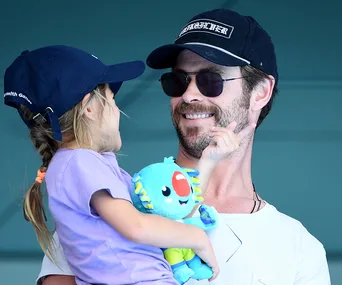 Father in cap and sunglasses holds daughter with stuffed toy at event, both smiling.