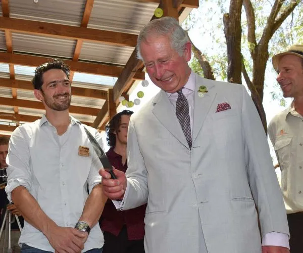 Prince Charles smiles while holding a knife, surrounded by three people in an outdoor setting.
