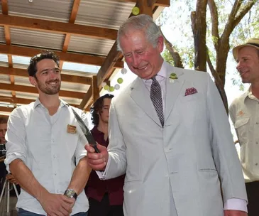 Prince Charles smiles while holding a knife, surrounded by three people in an outdoor setting.