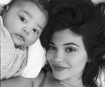 Two individuals lying on a bed, one is a baby and the other an adult woman with long dark hair, smiling. Black and white image.