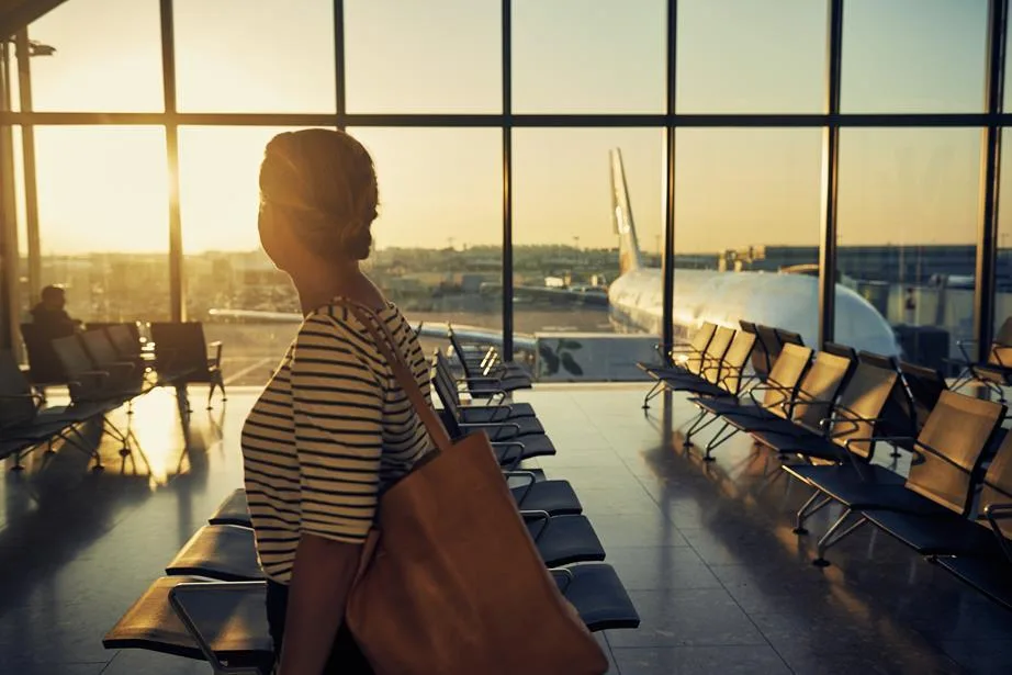 A traveler in a striped shirt stands in an airport terminal looking at a plane through large windows during sunset.