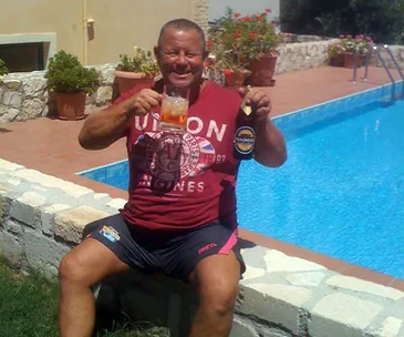 Man in red shirt sitting by a pool, holding a beer bottle and glass, smiling.