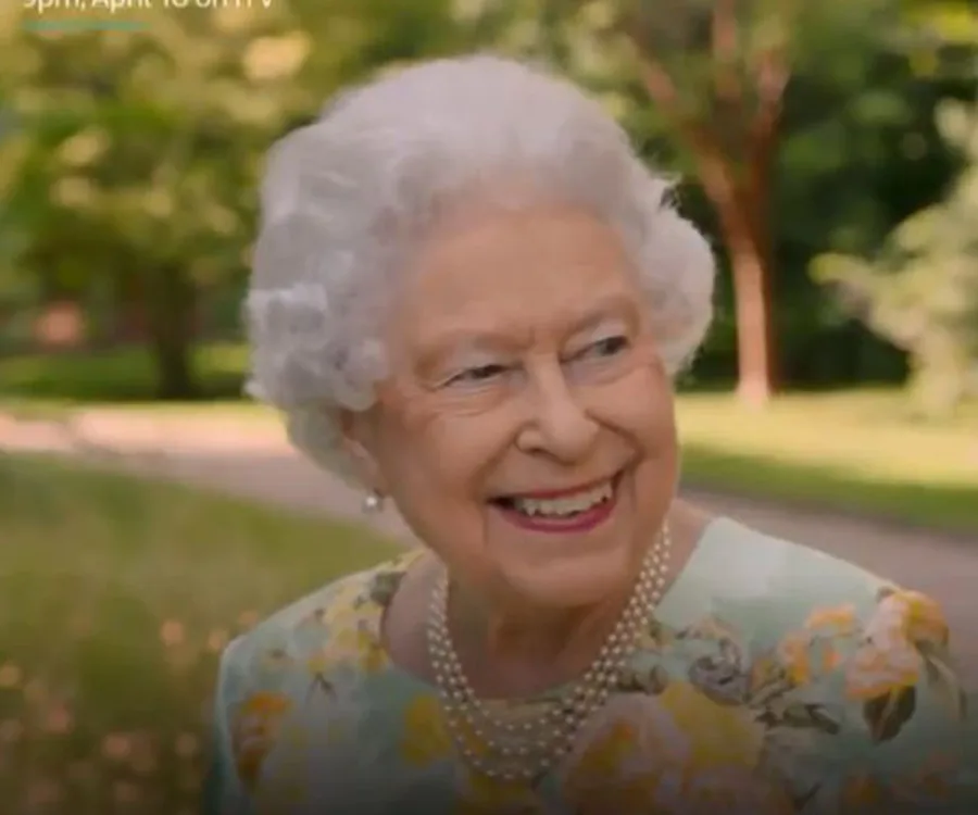 An elderly woman with white hair smiles while wearing a floral dress and pearl necklace in a garden setting.