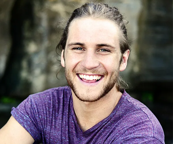 A smiling man with long hair and a beard, wearing a purple shirt, standing outdoors with a blurred natural background.