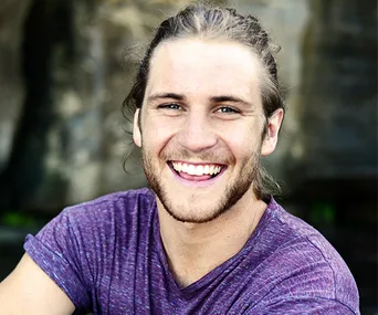 A smiling man with long hair and a beard, wearing a purple shirt, standing outdoors with a blurred natural background.