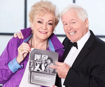 Two people smiling, holding a "Temptation" show photo. Both wear formal outfits, with the woman in purple and the man in a tuxedo.