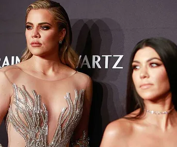 Two women at a formal event, one in a sheer embellished dress, the other blurred in the foreground, against a dark backdrop.