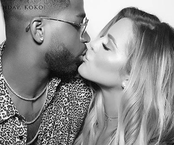 A couple shares a kiss in a grayscale photo; one wears glasses and a patterned shirt, the other has long hair.