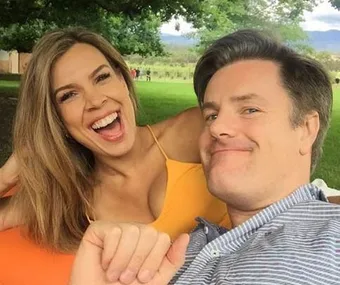 A laughing couple taking a selfie outdoors, sitting on a picnic blanket with trees in the background.
