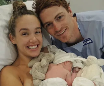 A smiling couple in a hospital holds newborn twins, with stuffed animals surrounding them.