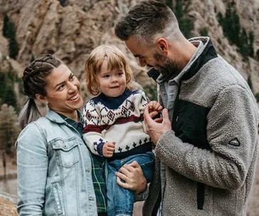 A couple smiling at their toddler, who is wearing a patterned sweater, outdoors with rocky scenery in the background.