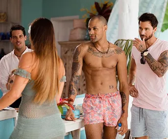 A shirtless man with tattoos and another in a pink shirt chat with a woman in a blue dress in a tropical setting.