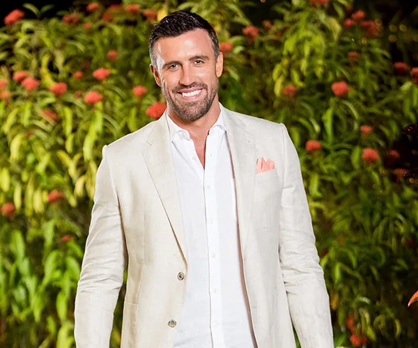 Man smiling in beige suit stands outdoors against lush green and floral background.
