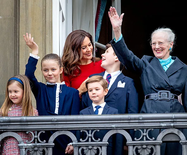 A family on a balcony waves; a woman in red and elderly woman in gray smile, with children in formal attire.