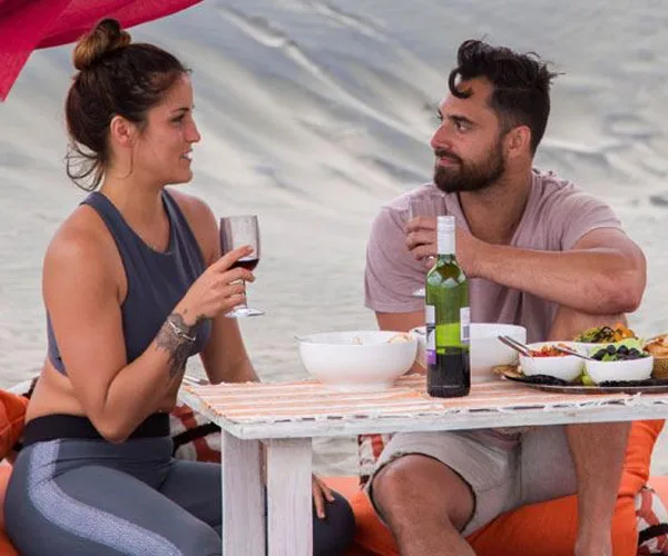 A couple enjoys wine and snacks at a beachside picnic table, engaged in conversation with a relaxed ambience.