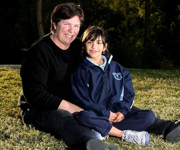 Two people sitting on grass, smiling. The adult wears black; the child in a school tracksuit. Outdoor, sunny setting.
