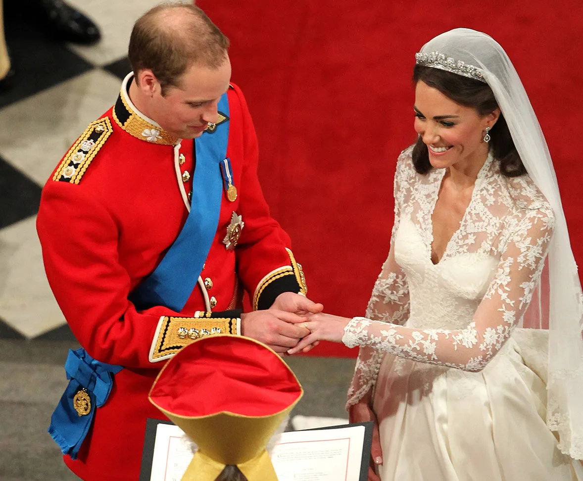 A bride in a lace gown and groom in a red military uniform exchange rings at a wedding ceremony.