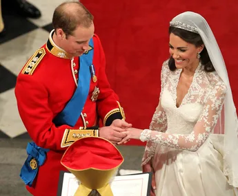 A bride in a lace gown and groom in a red military uniform exchange rings at a wedding ceremony.