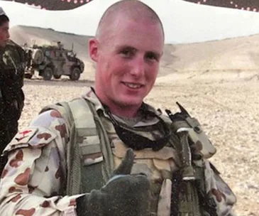 Soldier smiling and giving a thumbs-up in a desert setting with military vehicles in the background.