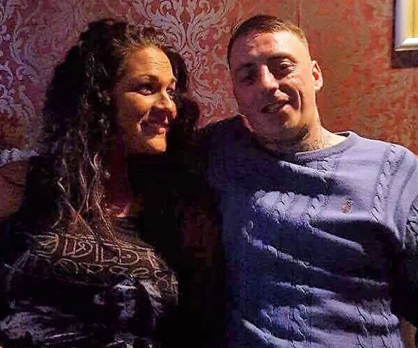 A couple sits together in a dimly lit room with patterned wallpaper, smiling at each other.