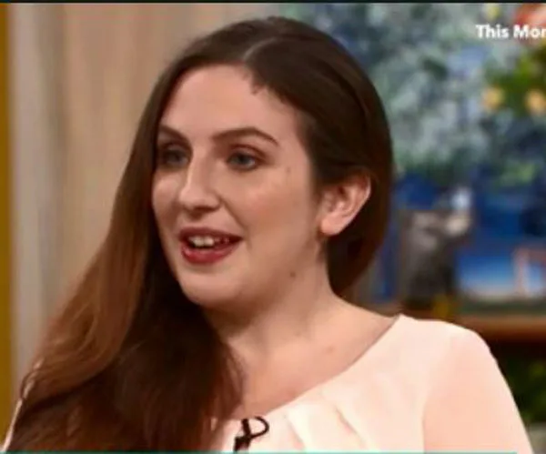 Woman with long brown hair smiles on a talk show set, wearing a light-colored top.