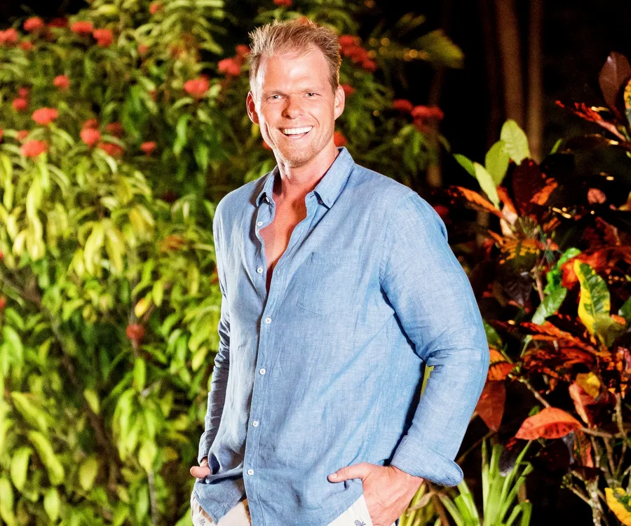 A smiling person in a blue shirt stands outdoors with green foliage and red flowers in the background.