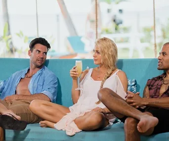 Three people relax on a blue sofa outdoors, one holding a drink, with tropical plants in the background.
