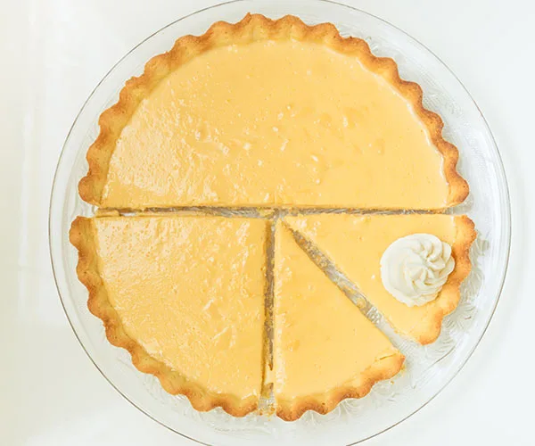 Lemon tart with a golden crust, sliced into six pieces, on a clear plate, garnished with a dollop of cream.