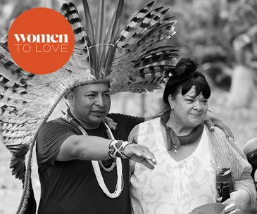 Two people, one wearing a feathered headdress, stand together outdoors in a "Women to Love" feature.