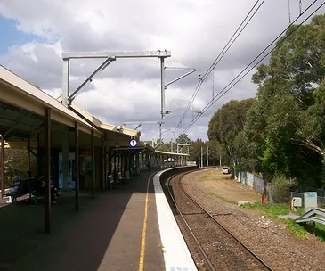 Sydney train station platform with tracks curving right, overhead power lines, and trees in the background.