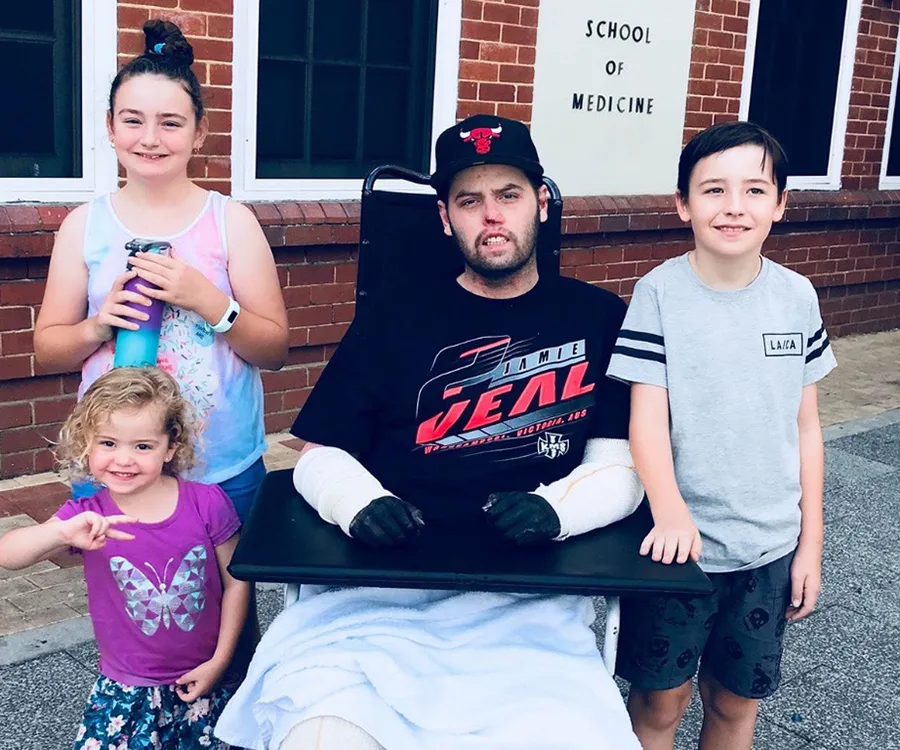 Man sitting in wheelchair with bandaged arms, surrounded by three children, outside a School of Medicine building.