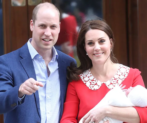 A couple smiling outside with a newborn baby in arms, with the woman in a red dress.