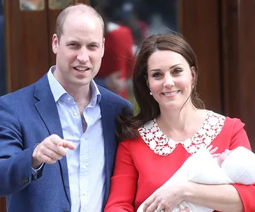 A couple smiling outside with a newborn baby in arms, with the woman in a red dress.