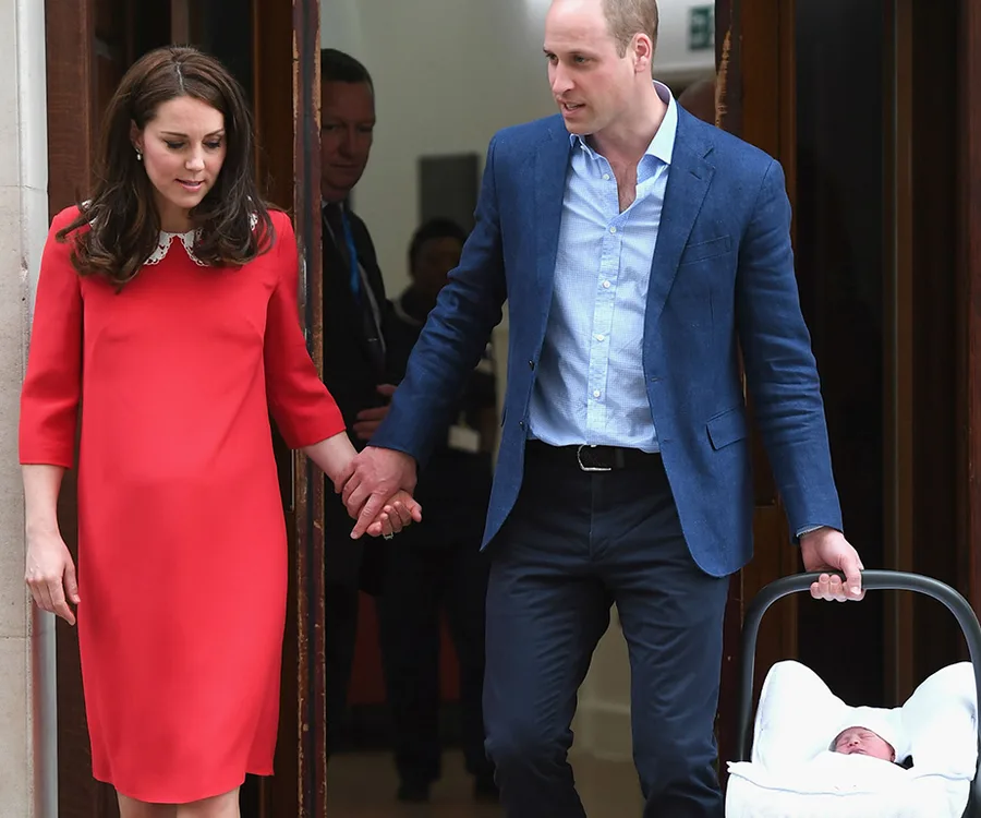 Royal couple exits a hospital holding hands, with a newborn in a carrier.