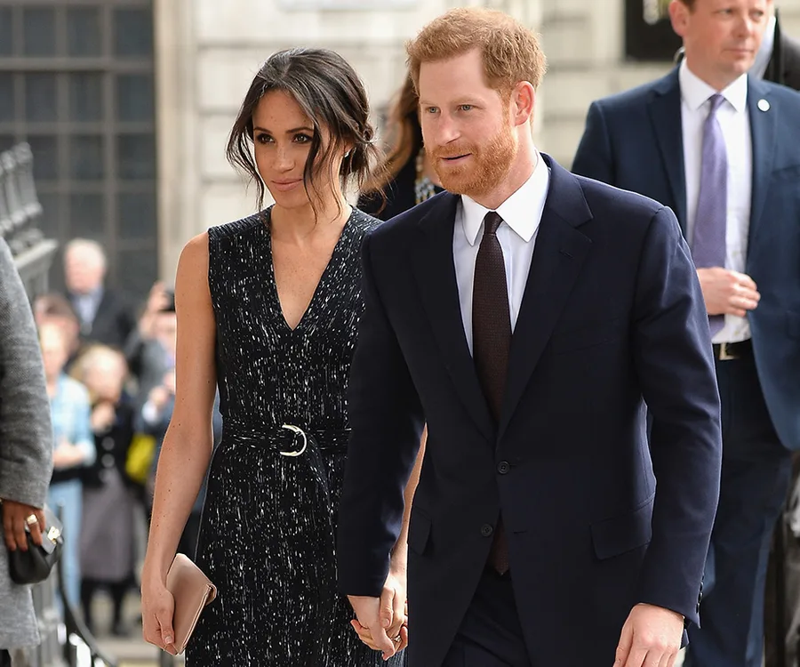 Couple walking hand in hand, dressed formally, outdoors with people in the background.
