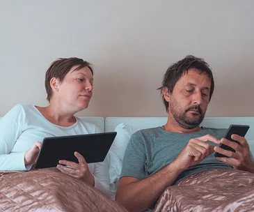 Couple in bed using a tablet and smartphone discreetly, looking focused on their devices.