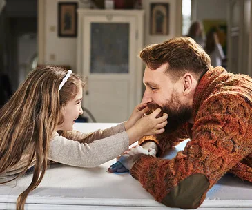 A young girl playfully holds a bearded man's face, both smiling warmly in a cozy room.