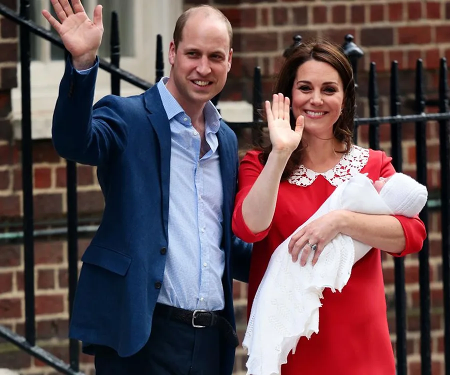 A couple waving outside a hospital, holding a newborn wrapped in a white blanket.