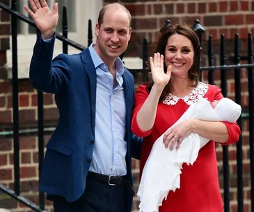 A couple waving outside a hospital, holding a newborn wrapped in a white blanket.
