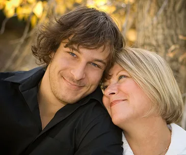 A mother and her adopted son share a warm, affectionate moment, smiling under a tree with autumn leaves in the background.