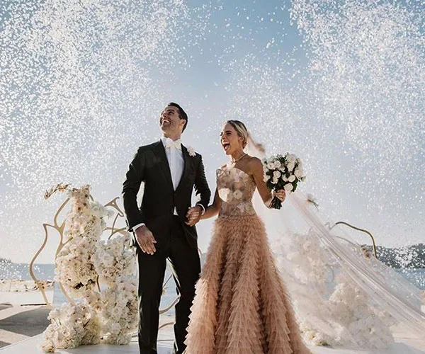 Bride in ruffled dress and groom in tux holding hands, surrounded by white flowers and confetti under a bright sky.