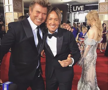 Two men in tuxedos smiling on the red carpet at an awards event, with a woman in a sequined dress nearby.