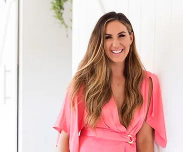 A smiling woman with long hair wearing a pink dress leans against a white wall indoors.