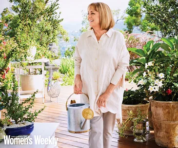 Woman smiling and holding a watering can on a lush garden deck, surrounded by potted plants and flowers.