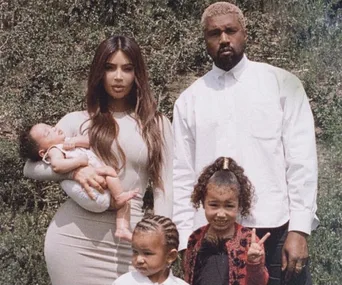 A family of five posing outdoors, with the mother holding a baby, and the father standing beside two young children.