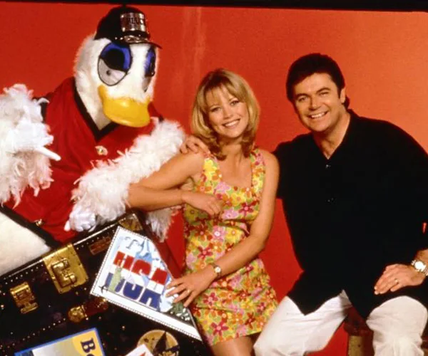 Three people, including someone in a duck costume, pose happily with a suitcase against an orange background.